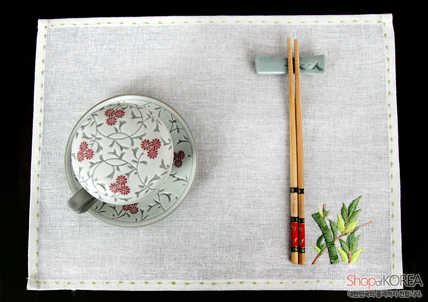 [텍스트: Shop of KOREA, 대한민국의 좋은 것까지 전합니다] 흰색 모시 식탁보 위 연두색 가장자리 스티치와 대나무 자수, 국화 문양 도자기와 나무 젓가락이 놓인 상차림 이미지