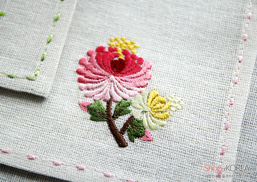 [텍스트: Shop of KOREA 대한민국의 품격까지 전합니다.] 옅은 회색 모시 위 분홍·노랑 꽃자수와 가장자리 스티치가 더해진 사군자 자수 식탁보 디테일 묘사