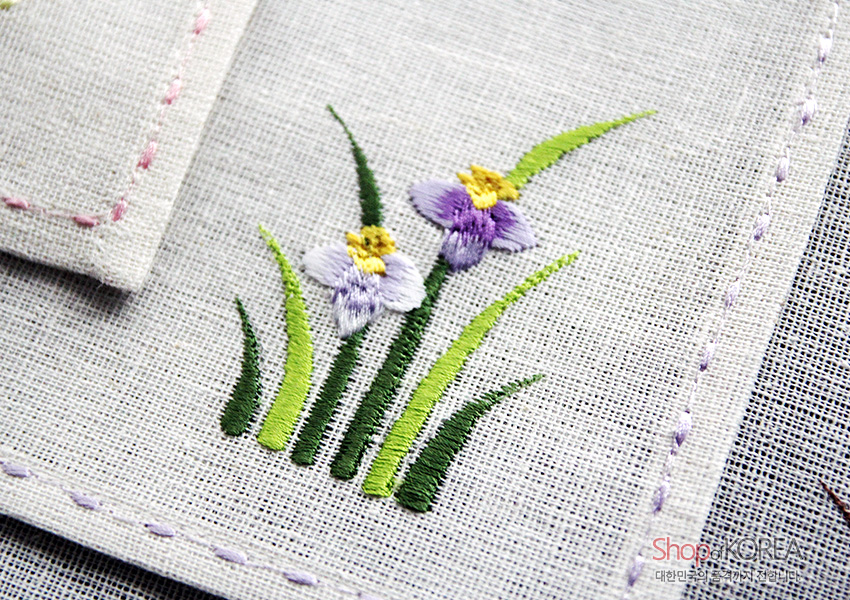 [텍스트: ShopKOREA 대한민국 문화의 즐거움까지 전합니다] 옅은 회색 모시 원단 위에 녹색 풀잎과 보라·노랑 색실로 수놓은 사군자 꽃 자수와 가장자리 스티치 장식 모습