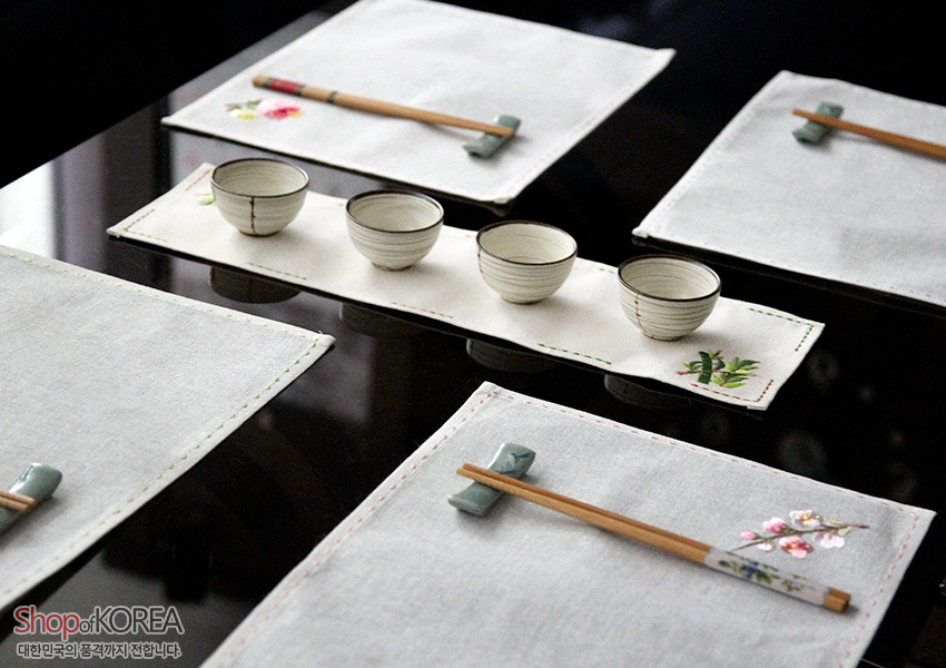 [텍스트: Shop of KOREA 대한민국의 품격까지 전합니다] 광목 재질 직사각 러너와 식탁매트, 끝단 흰꽃·초록 잎 자수와 가장자리 손누빔선 장식 식탁 구성 이미지