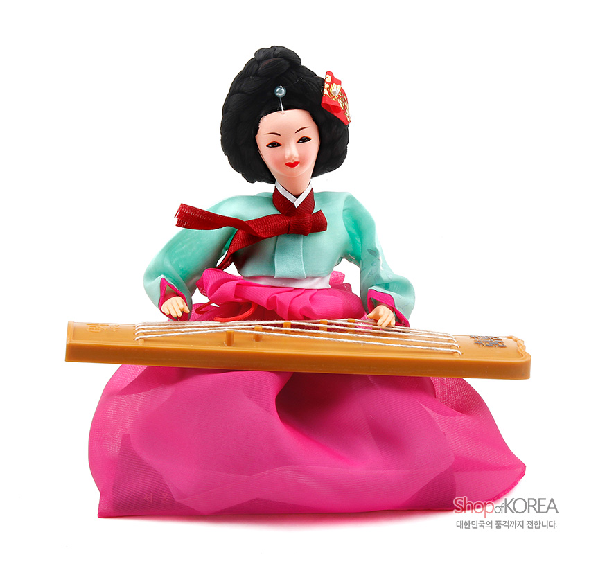 [텍스트: ShopofKOREA 대한민국의 품격까지 전합니다.] 비단 느낌 연두 저고리와 분홍색 겹겹 치마, 머리장식 더한 한복 인형이 나무 가야금을 연주하는 장식 모습