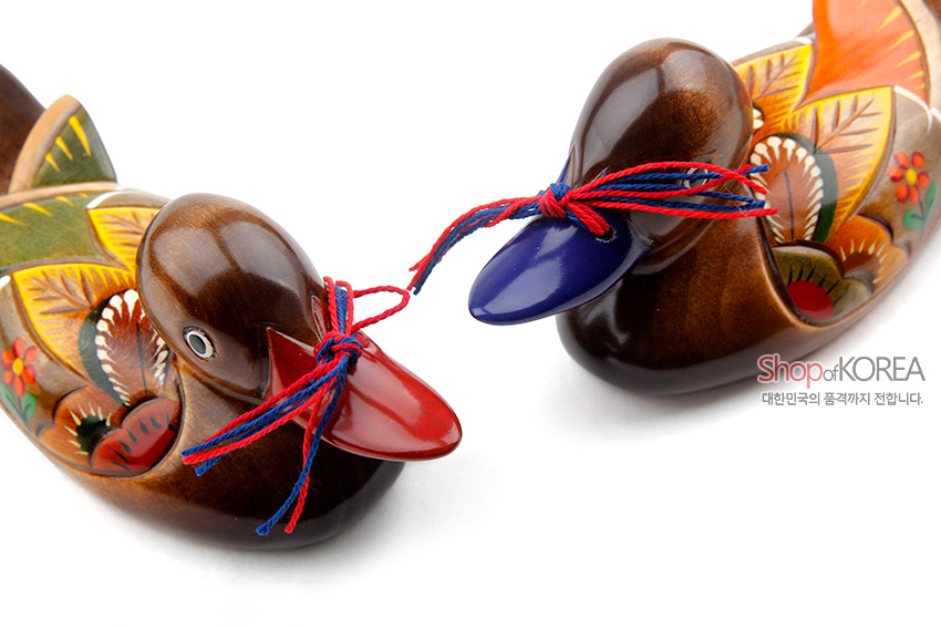 [텍스트: ShopofKOREA 대한민국의 품격까지 전합니다.] 옻칠 마감된 원목 원앙 한 쌍에 꽃과 잎 민화 문양을 채색하고 붉은색·파란색 오색 실 매듭을 더한 목각 장식품 이미지 설명