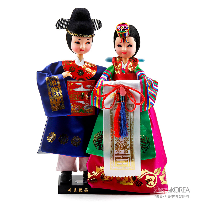 [텍스트: 서울民藝, ShopofKOREA 대한민국의 품격까지 전합니다.] 비단 소재 신랑신부 한복인형, 금박 봉황·연화 문양과 색동 소매, 매듭 장식이 돋보이는 전통 혼례복 재현 인형 세트 묘사