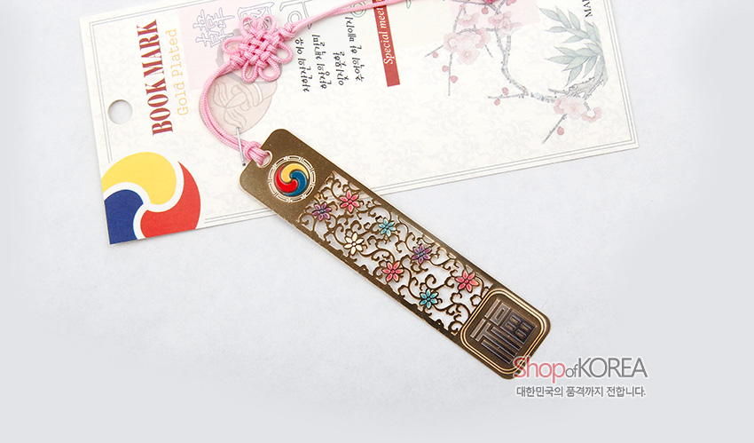 [텍스트: BOOKMARK, Gold Plated, shopofKOREA, 대한민국의 품격까지 전합니다] 금장 금속 바탕에 투각된 당초와 꽃문양, 상단 태극 문양과 분홍 전통 매듭 끈 장식 북마크 이미지