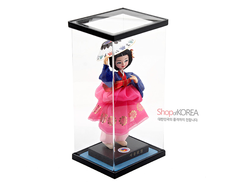 [텍스트: Shop of KOREA, 대한민국의 품격까지 전합니다., 서울 민속] 투명 아크릴 케이스 속 분홍 비단 치마에 금색 꽃문양 장식, 남색 저고리와 붉은 띠, 화려한 자수 양산을 든 전통 한복 인형 장식