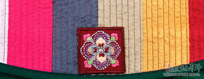 [텍스트: 샵오브코리아 대한민국 전통기념품 전문몰입니다] 적갈색 바탕의 누비색동 원단 위 중앙에 꽃문양 자수 패치가 더해진 디테일 장면, 분홍·회색·노랑·빨강 색동 배색과 녹색 가죽 마감선 대비 묘사