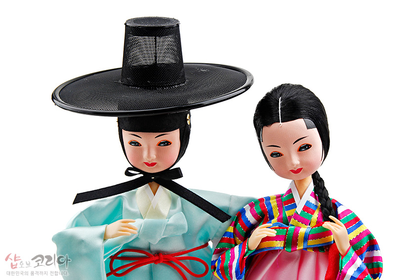 [텍스트: 샵오브코리아 대한민국의 품격까지 전해드립니다.] 비단 재질 한복을 입은 선비와 여인 인형, 흑색 갓과 옥색 도포, 오방색 줄무늬 저고리와 분홍 치마의 대비가 돋보이는 전통 의복 표현