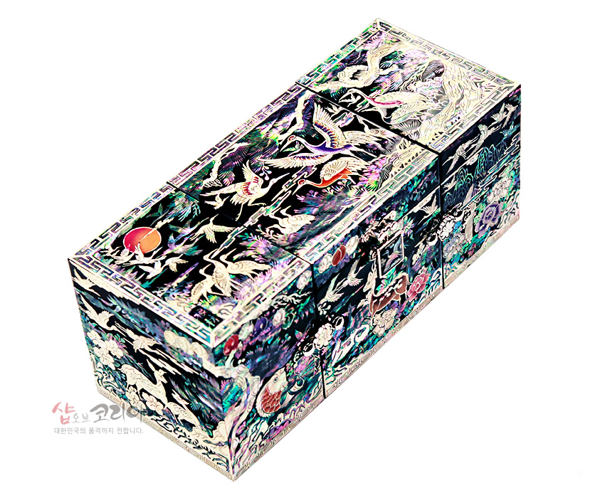 [텍스트: 샵오브코리아 대한민국의 품격까지 전합니다.] 흑색 옻칠 바탕 위에 다채로운 자개 상감으로 학, 해, 구름, 송죽과 장생도 문양을 정교하게 표현한 3폭 쌍합 보석함 모습