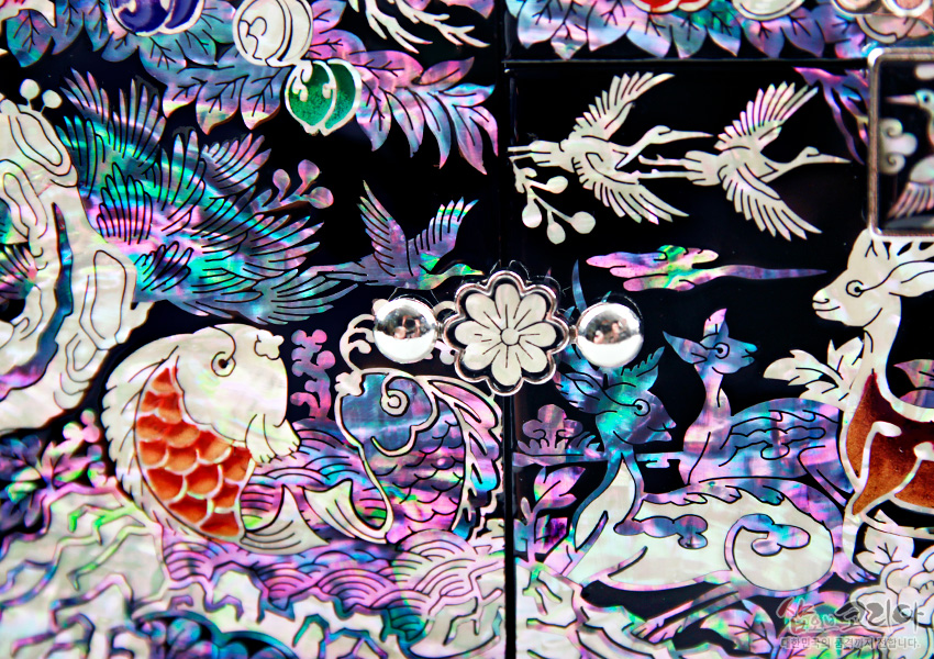 흑색 옻칠 바탕에 화려한 자개 상감으로 물고기와 학, 사슴, 파도와 구름, 수목이 어우러진 장생도 문양을 섬세하게 표현한 보석함 장식 면 구성