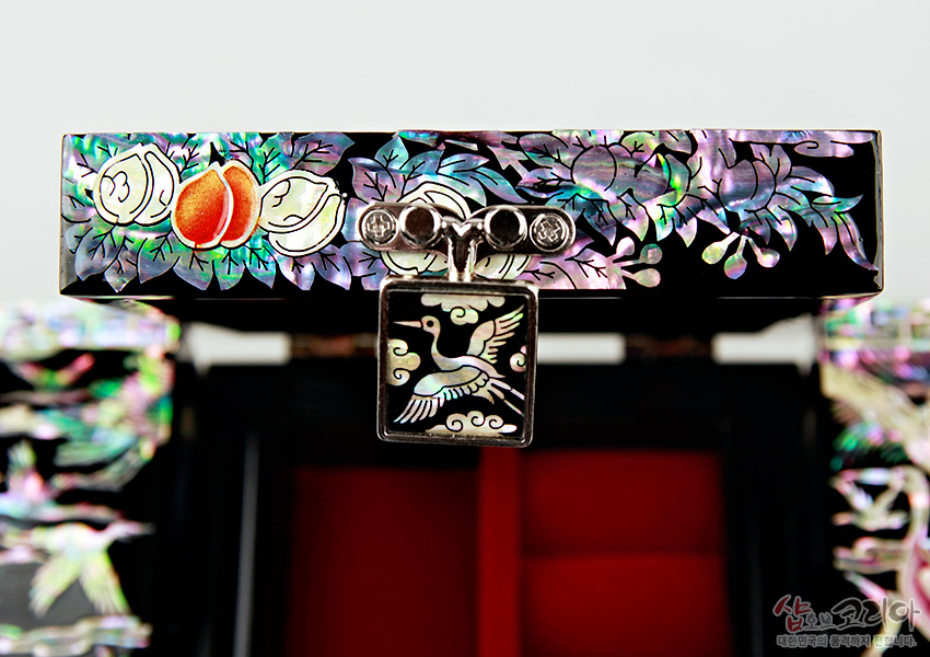 [텍스트: 샵오브코리아 대한민국의 품격까지 전합니다.] 흑색 옻칠 위 자개 상감 기법으로 복숭아와 잎사귀, 장생도 학 문양을 정교하게 표현한 잠금장치와 보석함 디테일 이미지