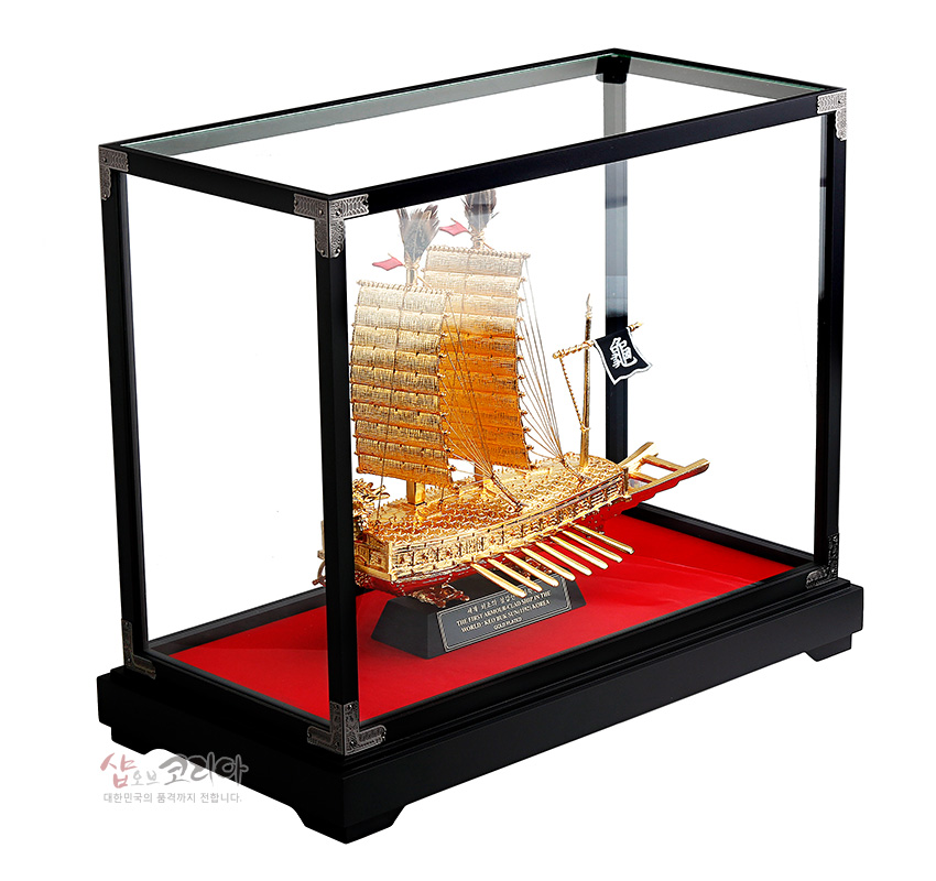 [텍스트: 龜] 금빛 금속 재질로 조각된 거북선과 돛, 노 장식을 유리 쇼케이스에 배치하고 검정 목재 프레임과 붉은 받침대로 대비를 준 구성