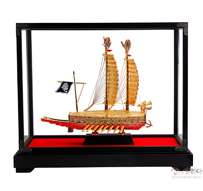 [텍스트: 龜] 금속 재질 24K 톤으로 표현된 용두와 갑판, 세밀한 노와 난간 장식, 검은 깃발과 붉은 받침대가 대비되는 거북선 모형 장식