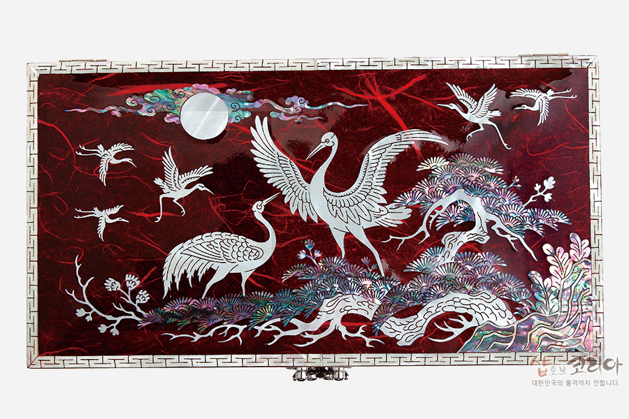 [텍스트: 샵오브코리아 대한민국의 품격까지 전합니다.] 붉은 옻칠 바탕 위에 자개 상감 기법으로 학과 소나무, 구름과 달, 파도 문양을 섬세하게 표현한 직사각형 보석함 상판 디자인