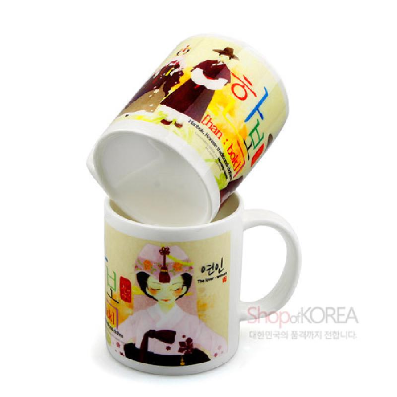 도자 재질의 머그컵에 한복 차림 남녀 일러스트와 한글 타이포그래피가 인쇄된 컬러 그래픽 디자인 제품.