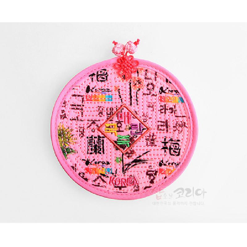 분홍색 천에 누비 기법으로 훈민정음 문자와 다채로운 전통 문양을 수놓은 원형 컵받침, 상단 노리개 매듭 장식.