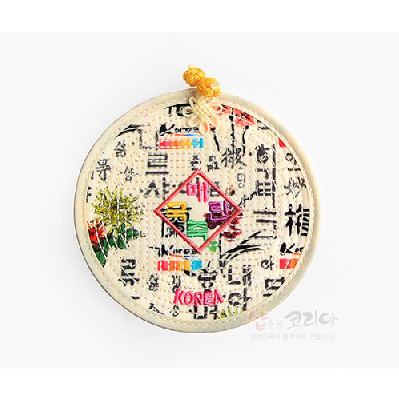 누빔 처리된 원형 천 재질에 훈민정음 서체와 오방색 자수 액자 문양, 매듭 장식이 어우러진 컵받침.