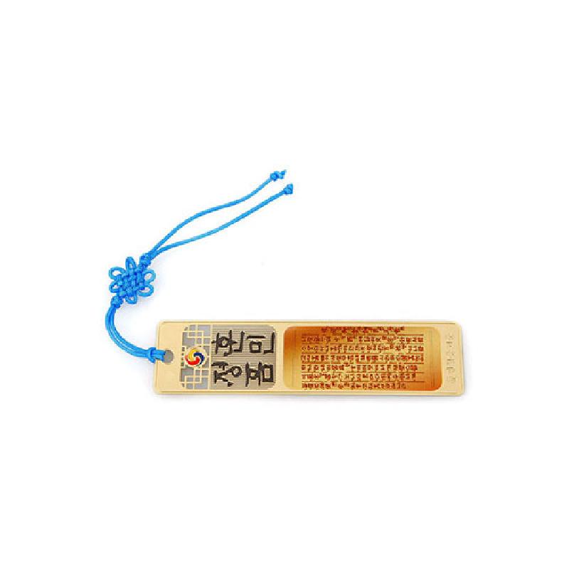 금속 재질에 금장 처리된 직사각형 책갈피, 훈민정음 한글 원문과 태극 문양, 청색 전통 매듭 끈 장식.