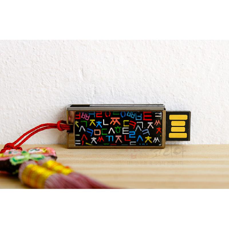 검은 바탕 위에 다채로운 훈민정음 자모 문양을 넣은 금속 USB와 붉은 끈 매듭 장식.