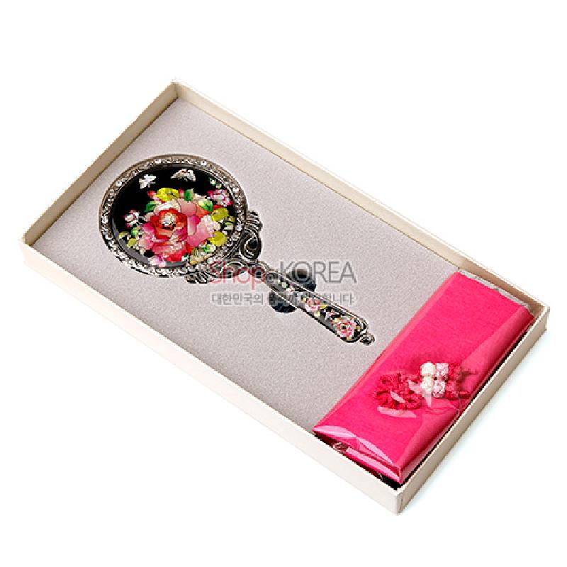 검은 바탕 위 자개 상감 장미와 나비 문양이 화려한 금속 손거울, 분홍 비단 주머니가 함께 구성된 세트.