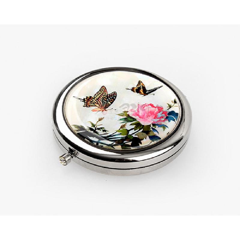 자개 상감 기법으로 장식된 원형 금속 손거울, 흰 바탕 위에 분홍 꽃과 나비 문양이 조화를 이룬 디자인.