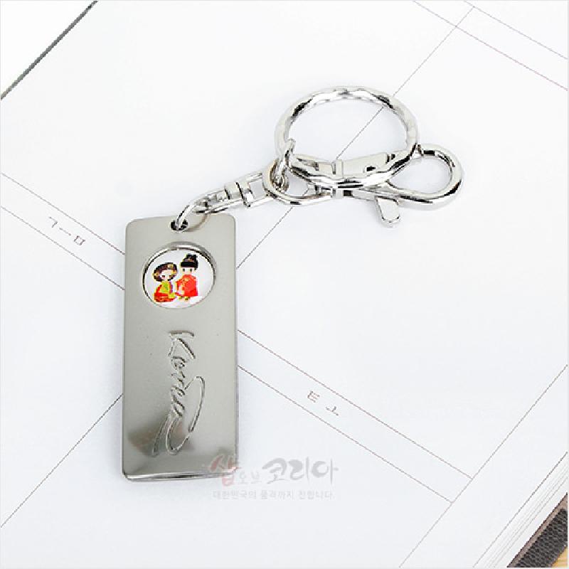 은색 금속 재질 직사각형 판에 한복 입은 남녀 캐릭터 원형 문양과 영문 각인이 들어간 열쇠고리. 은색 금속 재질 직사각형 판에 한복 입은 남녀 캐릭터 원형 문양과 영문 각인이 들어간 열쇠고리.
