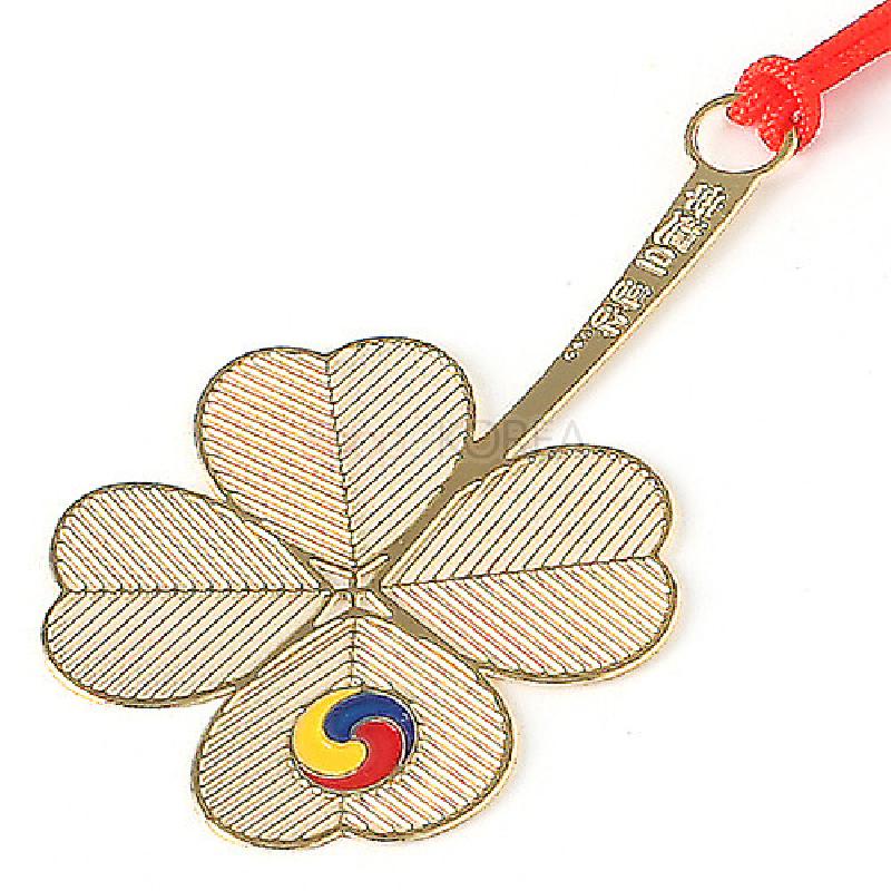 금속 재질의 네잎클로버 모양 책갈피, 잔줄 음각 기법과 태극 삼색 원형 문양, 상단 붉은 끈 장식.